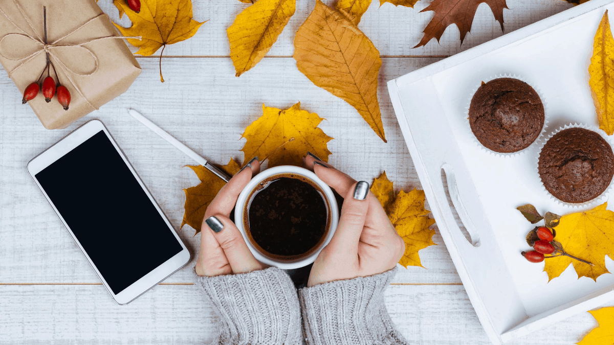 Fall Instagram captions - Women holding a cup of coffee on a home office table desk covered with fall leaves a gift and black screen smartphone 