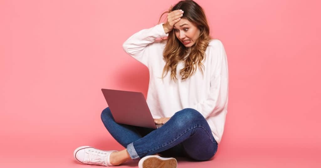 astonished young woman sitting with legs crossed, using laptop computer isolated over pink background